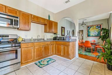 Bright open-plan kitchen with honey wood cabinets, granite countertops and stainless steel stove/microwave; tiled floor and tropical mat, arched doorway to a vibrant dining area with orange chairs, leafy rug, large tropical bird artwork and potted plants.