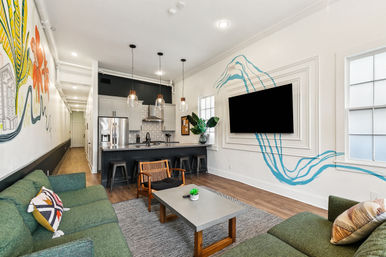 Open-plan urban apartment living room and kitchen with green sofas, concrete coffee table, wooden accent chair, black island with stools and pendant lights, stainless fridge, colorful mural and blue wave accent flowing around a wall-mounted TV.