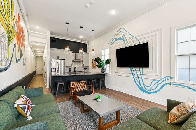 Open-plan urban apartment living room and kitchen with green sofas, concrete coffee table, wooden accent chair, black island with stools and pendant lights, stainless fridge, colorful mural and blue wave accent flowing around a wall-mounted TV.