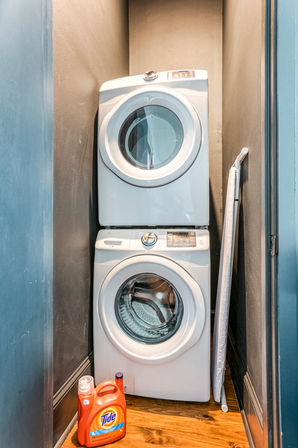 Stacked white front-loading washer and dryer in a narrow laundry closet, ironing board leaning against the wall and bright orange detergent jug on the wooden floor.