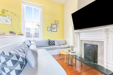 Bright sunny living room with a yellow accent wall, light-gray built-in sectional sofa topped with geometric throw pillows, hardwood floors, white Victorian-style mantel with black cast-iron fireplace and wall-mounted flat-screen TV, plus small yellow and green nested coffee tables, potted plants and framed art.