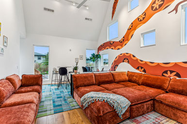 Spacious modern living room with rust-orange velvet sectional sofa, colorful patterned rugs, high vaulted ceiling, retro orange floral wall mural, multiple small windows and a dining nook with bar stools.