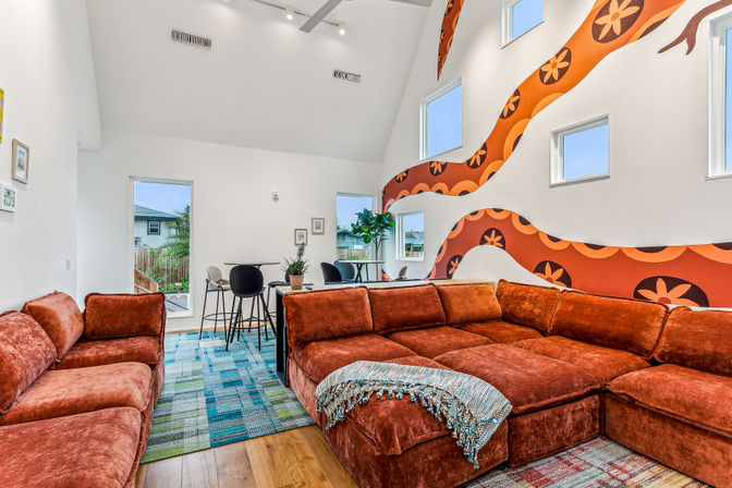 Spacious modern living room with rust-orange velvet sectional sofa, colorful patterned rugs, high vaulted ceiling, retro orange floral wall mural, multiple small windows and a dining nook with bar stools.