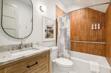 Bright modern home bathroom with wood vanity and stone countertop, round mirror, white toilet, bathtub with gray patterned shower curtain and warm terracotta tile surround with wall-mounted dispensers.