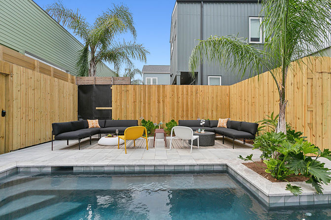 Modern urban backyard patio with a tiled plunge pool, black sectional sofas, yellow and white accent chairs, palm trees and wooden privacy fence.