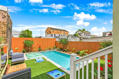 Bright urban backyard with a rectangular swimming pool, orange wooden privacy fence, artificial turf lounge area with gray outdoor sofas and cornhole boards, tropical palms and red flowers, neighboring houses under a vivid blue sky.