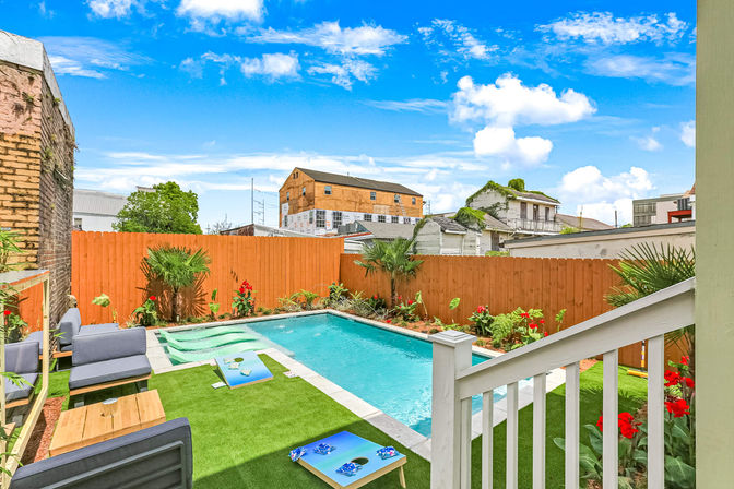 Bright urban backyard with a rectangular swimming pool, orange wooden privacy fence, artificial turf lounge area with gray outdoor sofas and cornhole boards, tropical palms and red flowers, neighboring houses under a vivid blue sky.