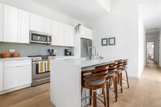 Bright modern white kitchen with waterfall quartz island and four curved wooden bar stools, stainless steel range and refrigerator, gray glass tile backsplash and light hardwood floors