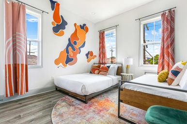 Bright modern twin bedroom with bold orange-and-navy abstract wall mural, two white beds with geometric throw pillows, coral patterned curtains, colorful round rug, wood-and-metal bed frames and sunlit windows.