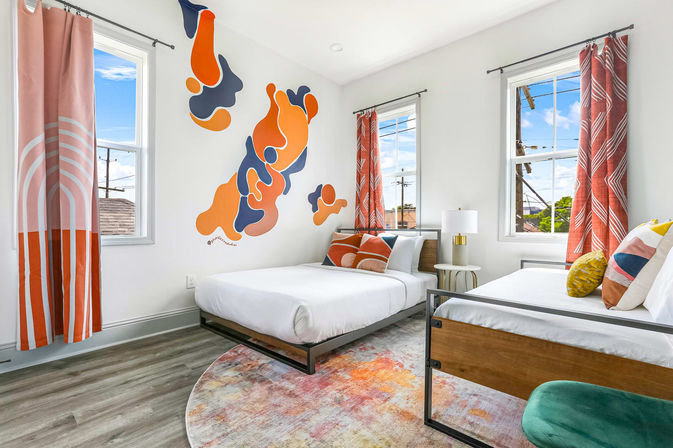 Bright modern twin bedroom with bold orange-and-navy abstract wall mural, two white beds with geometric throw pillows, coral patterned curtains, colorful round rug, wood-and-metal bed frames and sunlit windows.