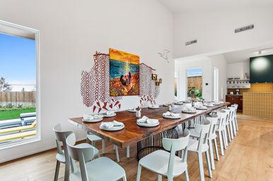 Sunlit modern dining room with a long reclaimed-wood communal table set for many, white minimalist chairs, colorful coastal wall art and decals, large window with backyard patio view and open-plan kitchen with wood floors.