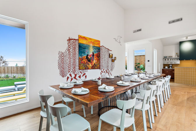 Sunlit modern dining room with a long reclaimed-wood communal table set for many, white minimalist chairs, colorful coastal wall art and decals, large window with backyard patio view and open-plan kitchen with wood floors.