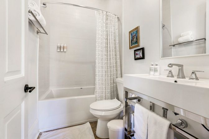 Bright white bathroom with tub-shower and patterned curtain, modern chrome sink and faucets, toilet, neatly folded towels and small framed wall art.