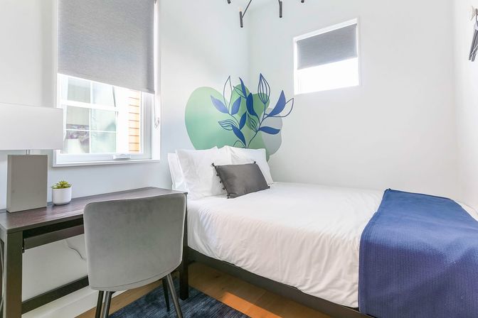 Bright, cozy modern apartment bedroom with a single bed dressed in white linens and a blue throw, gray accent pillow, leafy green wall mural behind the headboard, dark wood desk with lamp and small plant, and a gray velvet chair beneath windows with roller shades.
