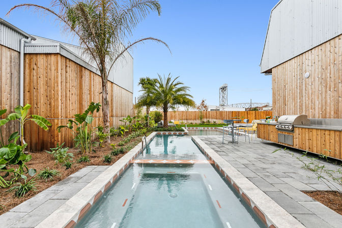 Sleek backyard lap pool with palm trees and tropical plants, wood-clad fencing and buildings, paved patio with stainless-steel outdoor grill and seating under a clear blue sky