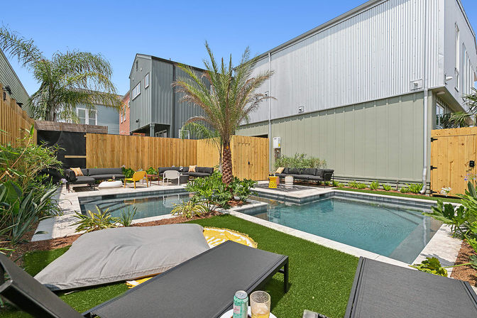 Sunny modern urban backyard oasis with a rectangular swimming pool and raised spa, central palm tree, artificial turf, black lounge chairs and sofas with yellow accent seating, wooden fence and metal-clad townhouses in the background.
