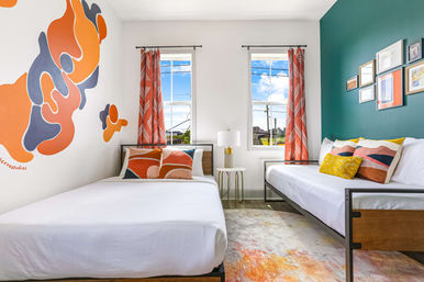 Bright modern twin guest room with teal accent wall and abstract orange mural, two beds with colorful pillows, patterned curtains and a bedside lamp