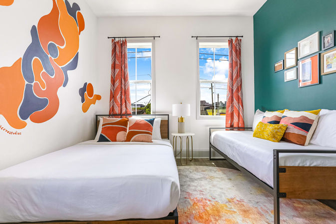 Bright modern twin guest room with teal accent wall and abstract orange mural, two beds with colorful pillows, patterned curtains and a bedside lamp