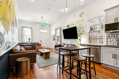 Bright urban apartment living room and kitchenette with hardwood floors, mural-accented white walls, leather sofas, wall-mounted TV, bar-height table with stools, pendant lights, and large windows.