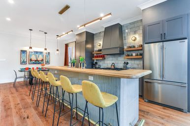 Bright modern open-plan kitchen with long wooden island and breakfast bar, yellow velvet bar stools, pendant lights, stainless steel refrigerator, and tiled backsplash