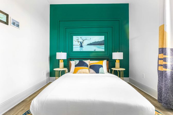 Bright modern bedroom with teal paneled feature wall, centered bed with white duvet, colorful pillows, gold bedside lamps and coastal artwork above the headboard.