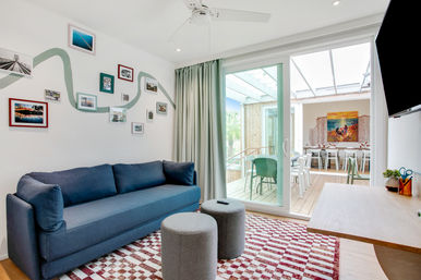 Bright modern living room with a navy sofa, gallery wall of framed photos, red-and-white checkered rug, two gray ottomans and sliding glass doors opening to a sunlit outdoor dining patio with green chairs.