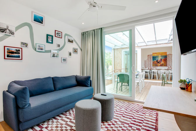 Bright modern living room with a navy sofa, gallery wall of framed photos, red-and-white checkered rug, two gray ottomans and sliding glass doors opening to a sunlit outdoor dining patio with green chairs.