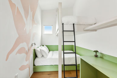Bright, cozy bunk bedroom with crisp white linens, green built-in lower bed and desk, black ladder to the top bunk, small square window, and a playful pink abstract wall mural.