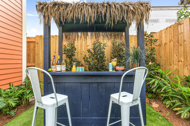 Home backyard tiki bar with thatched roof, navy wooden counter, two metal bar stools, tropical plants and colorful drinks on the bar.