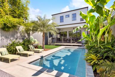 Sunny backyard rectangular pool at a modern two-story house with covered deck, palm tree, lush tropical plants and green lounge chairs