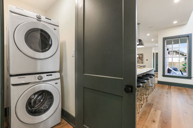 Stacked front-loading washer and dryer in a compact laundry closet opening to a modern open-plan kitchen with island, black bar stools, hardwood floors and a dark green door.