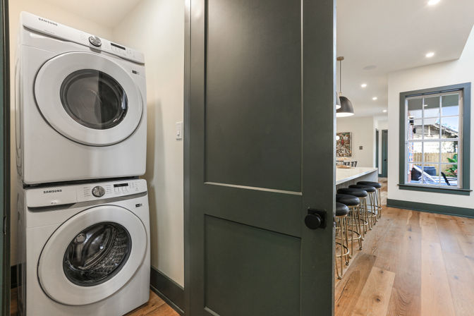Stacked front-loading washer and dryer in a compact laundry closet opening to a modern open-plan kitchen with island, black bar stools, hardwood floors and a dark green door.