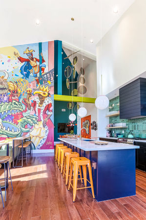 Bright high‑ceiling open kitchen with oversized colorful mural of whimsical carnival characters, navy island with white countertop, row of orange metal bar stools, globe pendant lights, green tile backsplash, and warm hardwood floors.