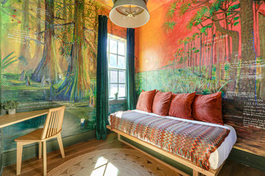 Cozy, sunlit guest room with a large hand-painted forest and wetland mural, daybed topped with rust-red corduroy pillows and a zigzag throw, wooden desk and chair by a window with deep green curtains, round jute rug and pendant light.
