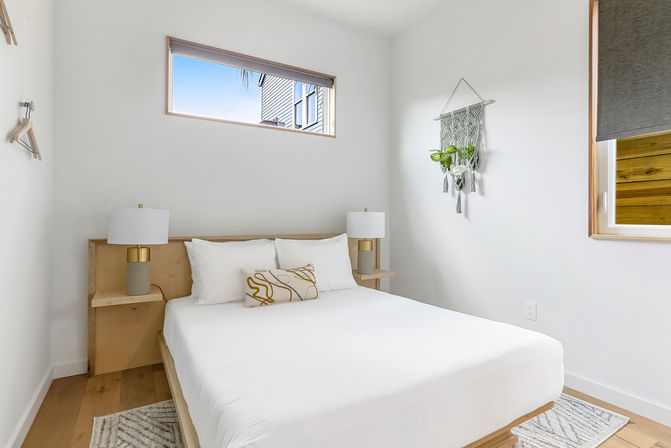 Bright minimalist bedroom with white walls and hardwood floors, wooden platform bed dressed in crisp white linens and a patterned pillow, built-in bedside shelves with gold-accent lamps, macramé plant hanger and narrow clerestory window letting in natural light.