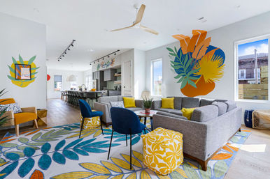 Sunny modern open-plan living room with a large tropical wall mural, gray sectional sofa, blue accent chairs, yellow pillows and patterned ottoman, botanical area rug, wood floors, ceiling fan, and a kitchen island visible in the background.