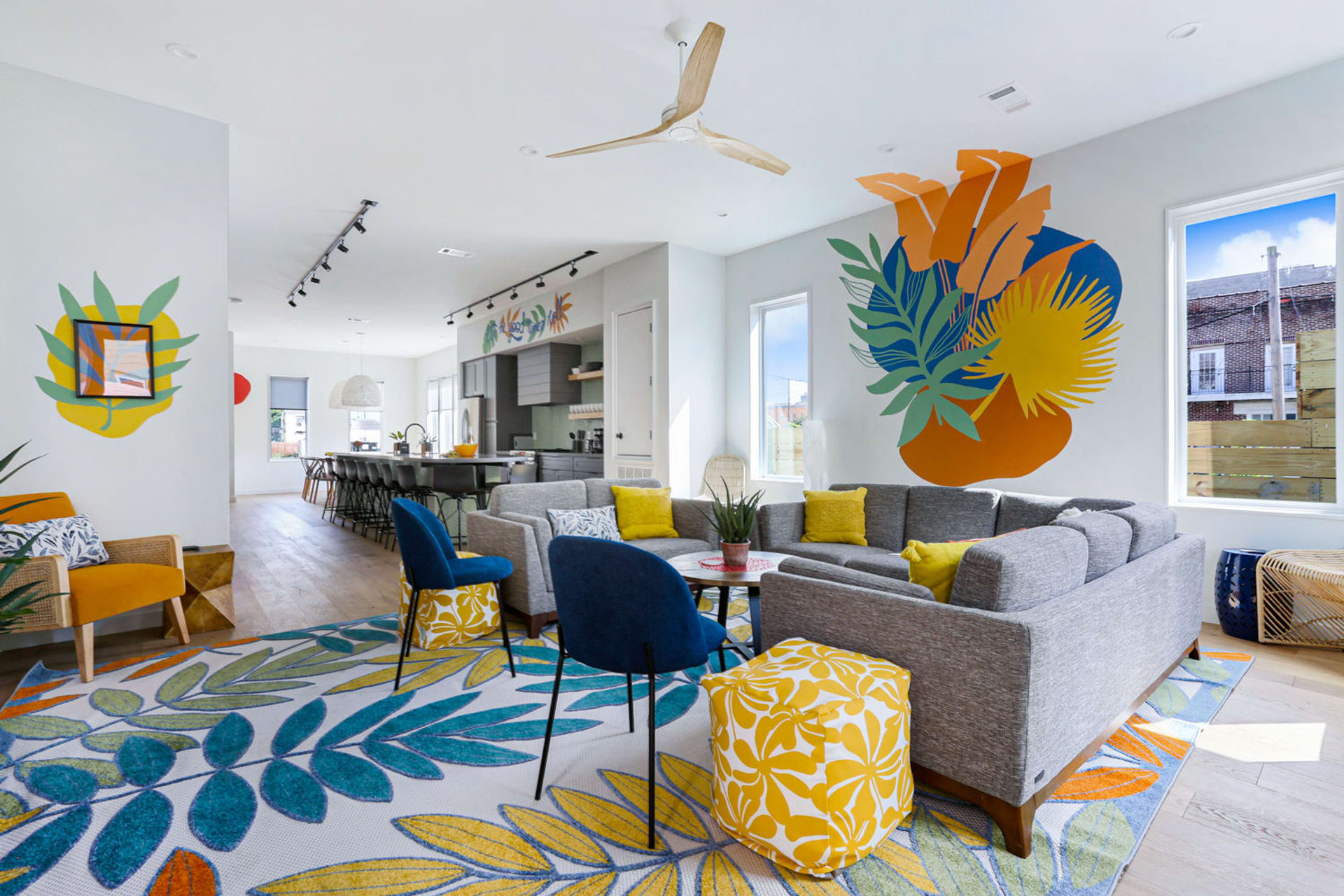 Sunny modern open-plan living room with a large tropical wall mural, gray sectional sofa, blue accent chairs, yellow pillows and patterned ottoman, botanical area rug, wood floors, ceiling fan, and a kitchen island visible in the background.