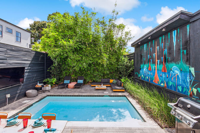 Modern urban backyard patio with rectangular turquoise pool, lounge chairs and sectional, tall bamboo privacy plants, colorful wall mural, outdoor dining table and stainless grill on a sunny day.