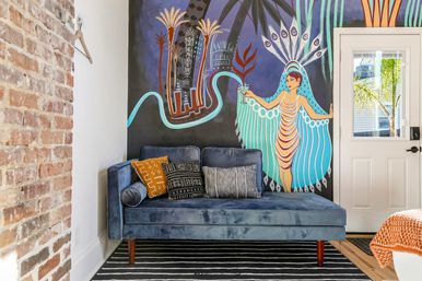 Blue velvet chaise sofa with patterned throw pillows against a colorful tropical mural of a dancing woman, exposed brick accent, striped rug and a white door with palm visible outside.