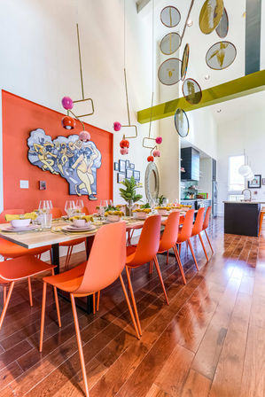 Vibrant modern dining room with long wooden table and row of orange chairs, colorful pendant lights, mural-accent wall, open-concept kitchen and glossy hardwood floors.