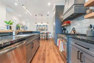 Modern open-plan kitchen and dining area with gray shaker cabinets, black speckled granite countertops, stainless steel appliances, hardwood floors, pendant lights, island sink, and a small dining table with colorful place settings and wall art.