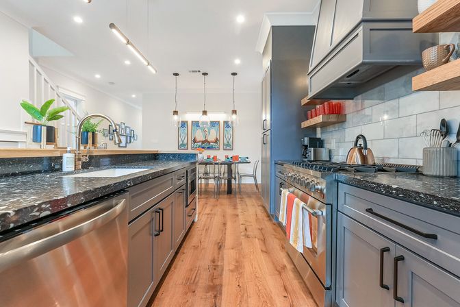 Modern open-plan kitchen and dining area with gray shaker cabinets, black speckled granite countertops, stainless steel appliances, hardwood floors, pendant lights, island sink, and a small dining table with colorful place settings and wall art.