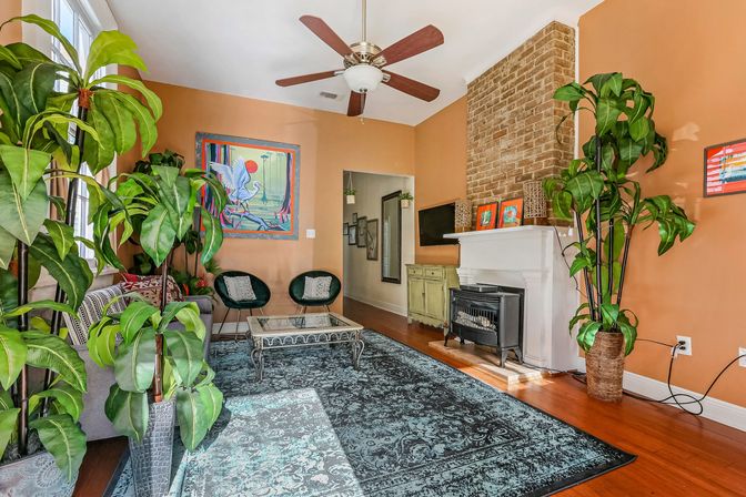 Sunlit eclectic living room with terracotta walls, exposed brick fireplace and white mantel, tall potted plants, teal patterned area rug, glass coffee table, two green accent chairs, colorful wall art and ceiling fan.