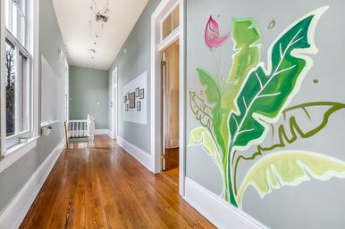 Sunlit upstairs hallway in a renovated home with warm hardwood floors, pale gray walls, framed gallery, large windows, and a bold tropical leaf mural beside an open doorway.