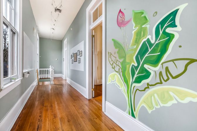 Sunlit upstairs hallway in a renovated home with warm hardwood floors, pale gray walls, framed gallery, large windows, and a bold tropical leaf mural beside an open doorway.