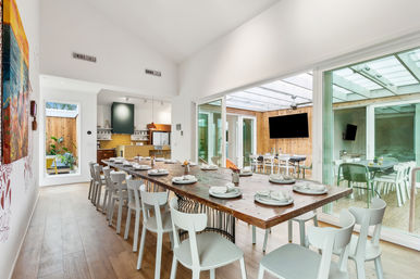 Bright modern dining area with high ceilings and a long rustic wooden communal table set for many, white chairs, open-plan kitchen in the background, and large sliding glass doors opening to a covered patio/sunroom with a wall-mounted TV.