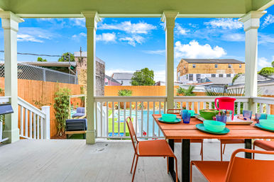 Covered residential porch overlooking a fenced backyard with a swimming pool and lawn, featuring a wooden outdoor dining table set with colorful bowls, glasses, a pitcher and orange chairs under a bright blue sky