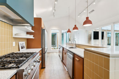 Bright modern open-concept kitchen with long peninsula island, quartz countertops, stainless steel gas range and fridge, warm terracotta cabinets, yellow tile backsplash, hardwood floors, and three red pendant lights over large windows