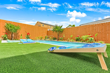 Sunny backyard with rectangular swimming pool, artificial turf lawn and wooden privacy fence, tropical plants and red flowers along the edge, two cornhole boards with blue patterned bean bags under a bright blue sky