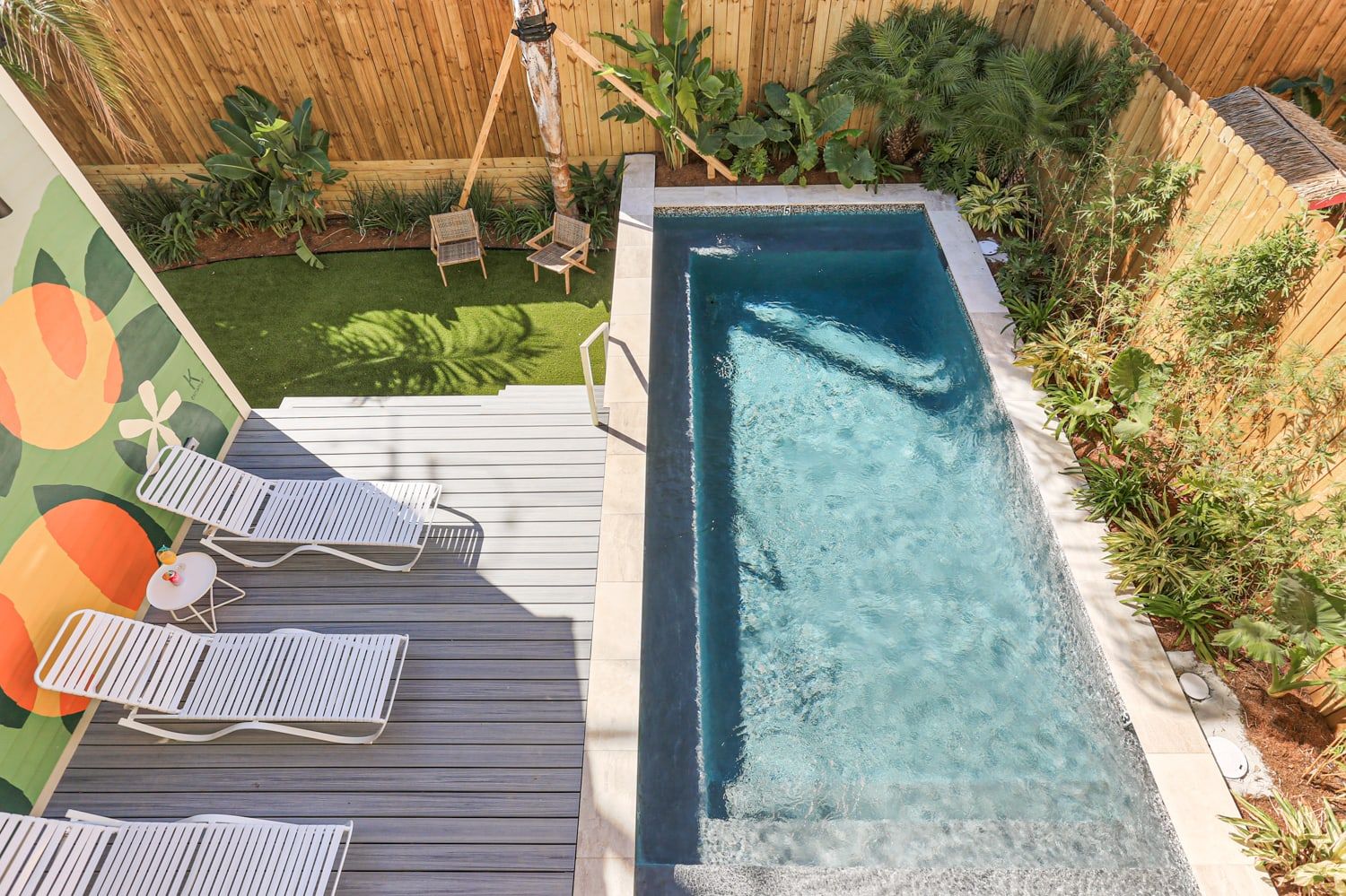 Aerial view of a rectangular backyard plunge pool with clear blue water, gray deck with white lounge chairs and small side table, colorful mural on the wall, and tropical plants along a wooden fence.
